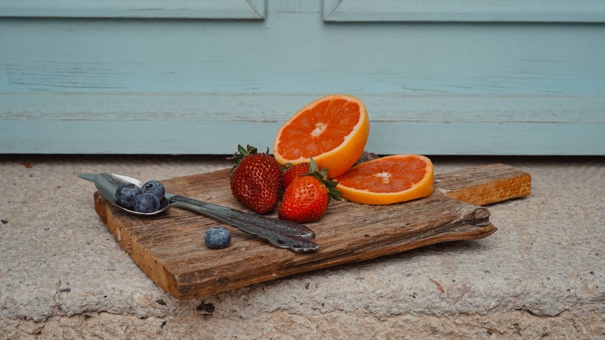 A rustic display of strawberries, oranges, and blueberries on a wooden board against a blue backdrop.